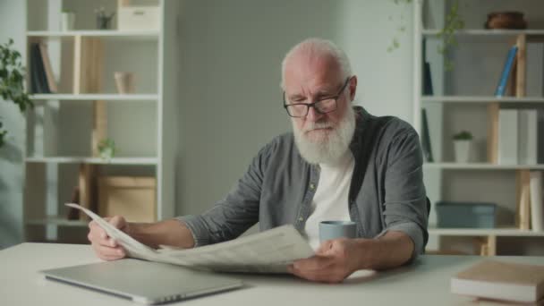一个严肃 聪明的老人坐在桌旁阅读报纸 一个拿着报纸的老年人 一个人在家喝咖啡 可以看到报纸上的新闻和时事 — 图库视频影像