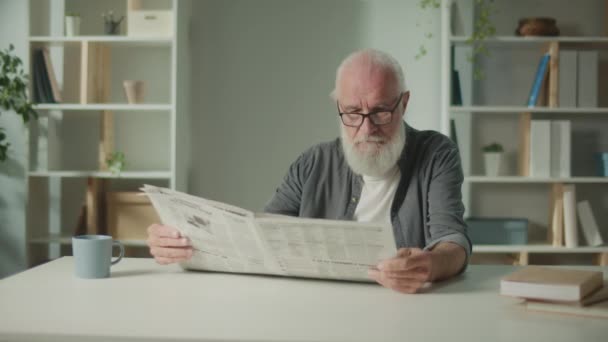 一位严肃 聪明的老人坐在桌旁读报 一位独自在家看报的老年人 在报纸上看到新闻和时事 — 图库视频影像