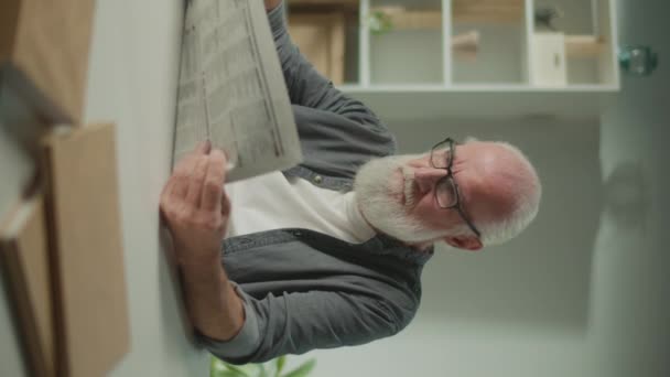 一个严肃 聪明的老人坐在桌旁看报纸 一个老年人一个人在家看报纸 就能看到报纸上的新闻和时事 — 图库视频影像