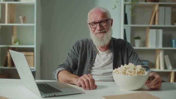ノートパソコンとポップコーンとメガネの笑顔の老人の肖像画 陽気な老人は面白い映画を見に行きます リラクゼーションと心理的救済の形としての映画 — ストック動画