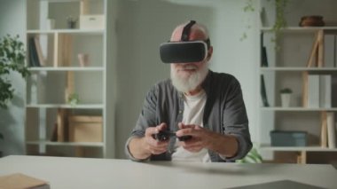 VR gözlüklü yaşlı bir adam, oyun çubuğuyla bilgisayar oyunu oynuyor masada oturuyor. Modern bir yaşlı adam PlayStation oynuyor. Yaşlılar için Artırılmış Gerçeklik ve Oyun.