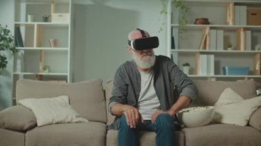 VR gözlüklü, gri sakallı, modern bir yaşlı adam çevredeki her şeyi inceler. VR gözlüklü, elinde patlamış mısırla koltukta oturan yaşlı bir adam. Yaşlılara Yardım İçin VR.