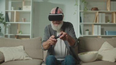 VR gözlüklü yaşlı bir adam, Joystick ile bilgisayar oyunu oynar, koltukta oturur ve mısır yer. Modern yaşlı bir adam PlayStation oynar. Yaşlılar için Artırılmış Gerçeklik ve Oyun.