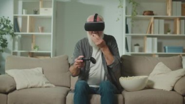 VR gözlüklü yaşlı bir adam, Joystick ile bilgisayar oyunu oynar, koltukta oturur ve mısır yer. Modern yaşlı bir adam PlayStation oynar. Yaşlılar için Artırılmış Gerçeklik ve Oyun.
