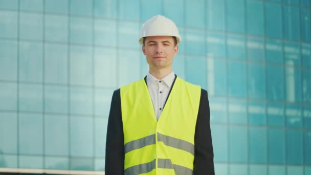 成功した若い男性エンジニアまたはヘルメットと安全ジャケットで身に着けている建築家の肖像画 青いWindowsの建物の近くに立っている文書を保持 — ストック動画
