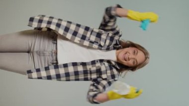 Dikey Bakış. Sarı Eldiven Giyen Mutlu Genç Bir Kadının Portresi, Elinde Temizleyici Sprey Şişe ve Paçavra, Gri Arkaplanda Temizlik Başarısı 'nda Neşeleniyor. Akıllı Temizlik Teknolojisi.
