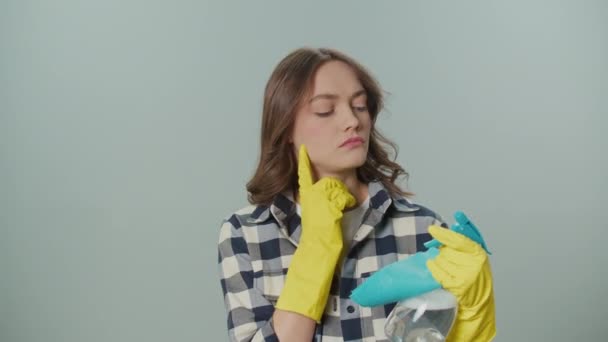 一个穿着黄色手套 头脑清醒的年轻女人的画像 她拿着一个清洁喷雾器 在灰色的背景上擦着碎布 一位女家庭主妇想从哪里开始清洁 卫生和安全议定书 — 图库视频影像