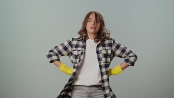疲れた若い女性は黄色い保護ゴム製手袋を身に着け 彼女の側面に彼女の手を保ち 灰色の背景の仕事のクリーニングの量を調査します デジタル用具が付いているスマートなクリーニング — ストック動画