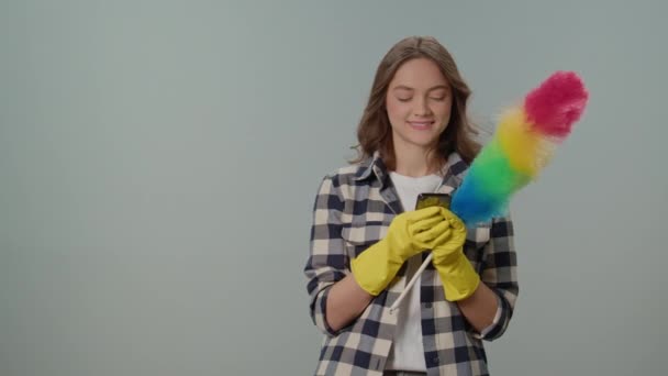 一位戴着黄色手套 头戴垃圾箱的快乐的年轻女性 在灰色背景下通过电话获得成功 她为此感到高兴 无线和应用控制的清洁工具 — 图库视频影像