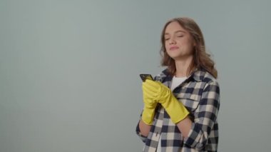 Sarı Eldivenli Gülümseyen Genç Bir Kadın, Gri Arkaplanda, Akıllı Telefonuyla Mesajlaşıyor. Sanal Temizlik Danışmanlıkları.
