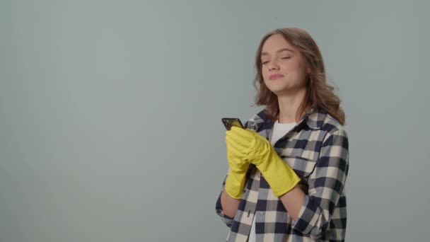 一位穿着黄色手套 面带微笑的年轻女性家庭主妇在灰色背景的智能手机上与人发短信 虚拟清洁咨询 — 图库视频影像