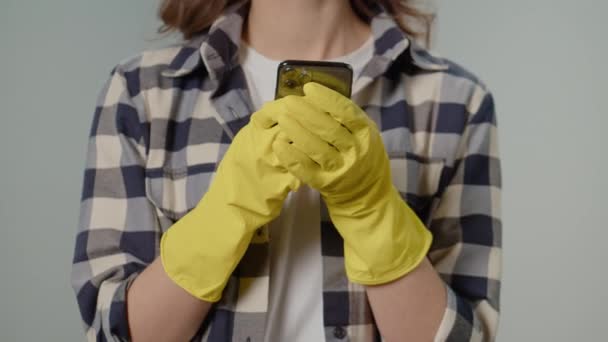 戴黄色手套的女性 中的智能手机特写 一位穿着黄色手套的年轻女性家庭主妇在灰蒙蒙的背景下 用智能手机与人发短信 虚拟清洁咨询 — 图库视频影像