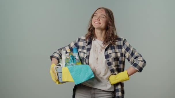 一位穿着黄色橡胶护目镜 拿着一个装有清洁用品的盒子 在灰色背景上对自己的清洁卫生结果感到满意的微笑的肮脏的年轻女人 — 图库视频影像