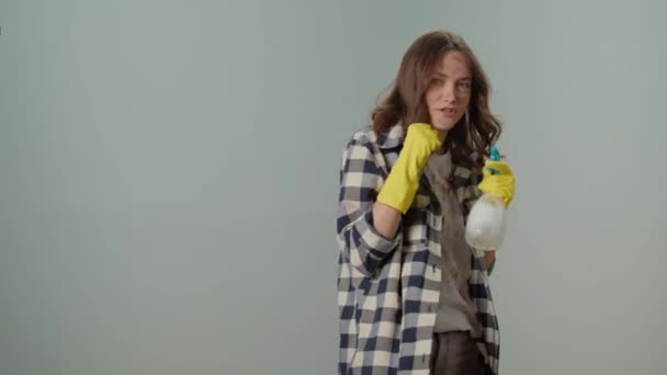 肮脏而严肃的年轻女性家庭主妇 戴着黄色手套 手里拿着一个清洁喷雾器和一把破布 灰色背景上的污迹斑斑 多功能电器的简约清洁 — 图库视频影像