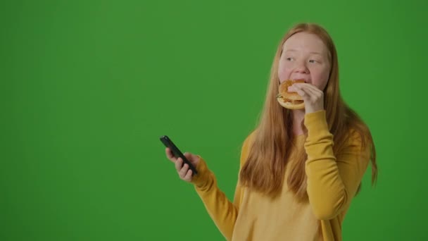 垂直ビデオ グリーン スクリーン ガールは同時にバーガーを食べながら携帯電話でブラウズする 現代のマルチタスクのスナップショットとファストフード文化とのデジタルライフの融合 — ストック動画