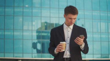 Kahve molası sırasında resmi takım elbiseli genç bir erkek mühendis veya mimar mavi pencereli bir binanın yanında duruyor ve telefonla konuşuyor.