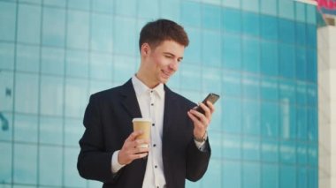 Kahve molası sırasında resmi takım elbiseli genç bir erkek mühendis veya mimar mavi pencereli bir binanın yanında duruyor ve telefonla konuşuyor.