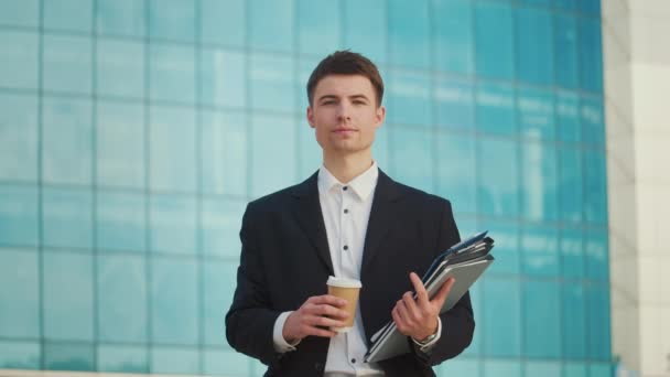 成功した若い男性エンジニアまたは建築家がブルーウィンドウで建物の近くに立っているドキュメントを着用している肖像画 — ストック動画