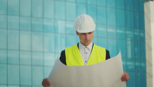 ヘルメットおよび安全ジャケットで身に着けている男性エンジニアまたは建築家青い窓が付いている建物の近くで歩く彼の手の構造図を見て — ストック動画