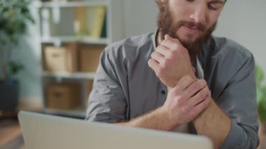 Bilek ağrısı. Bir adam laptopta çalışırken aniden bileğinde ağrı hissetti. Yaralı koluna diğer eliyle dokunuyor. Ofis sendromu konsepti. Klavye Kullanmaktan El Ağrısı.