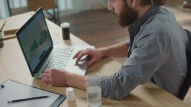 Bilek ağrısı. Bir adam laptopta çalışırken aniden bileğinde ağrı hissetti. Yaralı koluna diğer eliyle dokunuyor. Ofis sendromu konsepti. Klavye Kullanmaktan El Ağrısı.