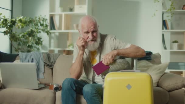 一个带着护照和手提箱的令人失望的老人旅行者 一个上了年纪的男人不喜欢不成功的旅行 坐在沙发上 护照问题 旅行规划 — 图库视频影像