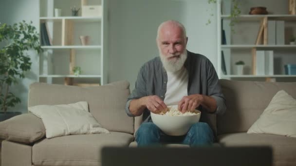 老人はポップコーンを食べ スポーツプログラムを見ます ソファーに座ってスポーツの勝利を祝います リラクゼーションと心理的救済の形としての映画 — ストック動画