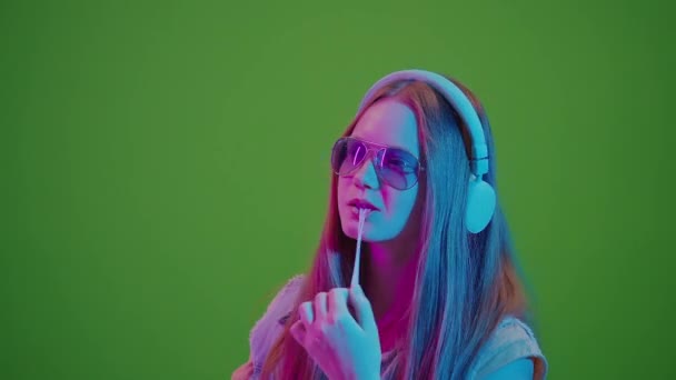 绿色屏风 少女在霓虹灯中跳舞 沉浸在耳机里的音乐中 咀嚼着一大杯粉红泡泡糖 复古情调与现代青年文化的融合与对音乐的热爱 — 图库视频影像