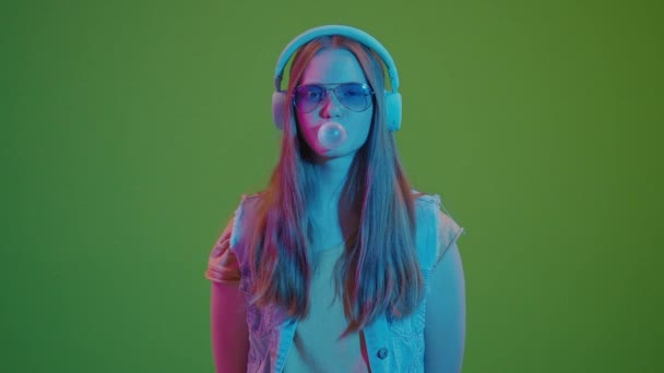 グリーン スクリーン ネオンライトで踊るティーンエイジャーガール 彼女のヘッドフォンから音楽に没頭 大きなピンクバブルガムを噛みます 現代の若者文化と音楽への情熱のレトロバイブの融合 — ストック動画