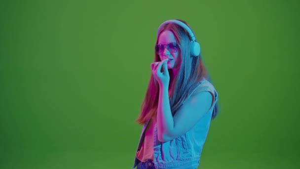 グリーン スクリーン ネオンライトで踊るティーンエイジャーガール 彼女のヘッドフォンから音楽に没頭 大きなピンクバブルガムを噛みます 現代の若者文化と音楽への情熱のレトロバイブの融合 — ストック動画