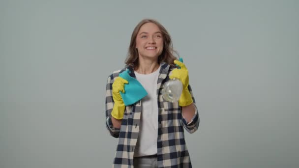 一位穿着黄色手套的微笑的年轻女子的画像 手里拿着一个洁净的喷雾器和碎布 在灰色的背景上跳舞 一位女家庭主妇正准备清扫 剥落的黑客 — 图库视频影像