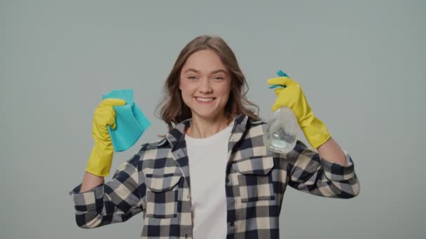 一位穿着黄色手套的微笑的年轻女子的画像 手里拿着一个洁净的喷雾器和碎布 在灰色的背景上跳舞 一位女家庭主妇正准备清扫 速度清洗 — 图库视频影像