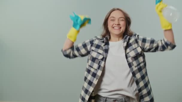 一个穿着黄色手套的快乐的年轻女人的画像 拿着一个清洁的喷雾器和碎布 为清洁的成功感到高兴 在灰色的背景上欢蹦乱跳 — 图库视频影像