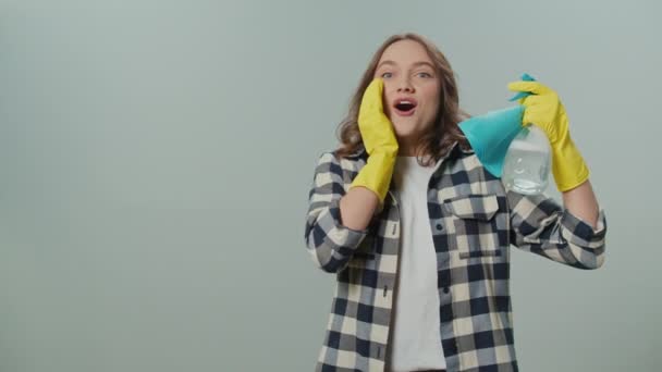 黄色い手袋の驚くべき若い女性の肖像画 グレイバックグラウンドにクリーニングスプレーボトルとラグを置きます 女性主婦が掃除する準備をしています 多機能クリーニングツール — ストック動画