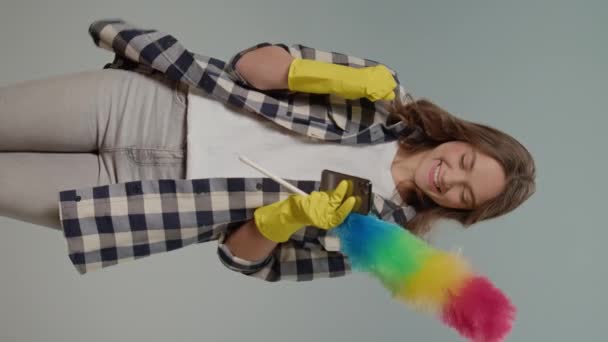 垂直ビュー 黄色い手袋とダストパンで幸せな若い女性 グレイバックグラウンドの電話で成功する喜び 無線およびアプリケーション制御のクリーニングアプライアンス — ストック動画