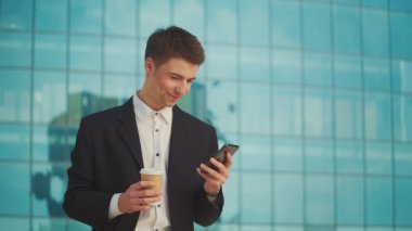 Kahve molası sırasında resmi takım elbiseli genç bir erkek mühendis veya mimar mavi pencereli bir binanın yanında duruyor ve telefonla konuşuyor.
