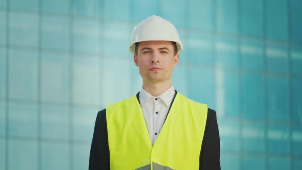 一名成功的年轻男性工程师或穿着头盔和安全夹克的建筑师的画像 他拿着站在蓝窗大楼附近的文件 — 图库视频影像