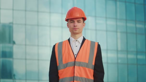 成功した若い男性エンジニアまたはヘルメットと安全ジャケットで身に着けている建築家の肖像画 青いWindowsの建物の近くに立っている文書を保持 — ストック動画