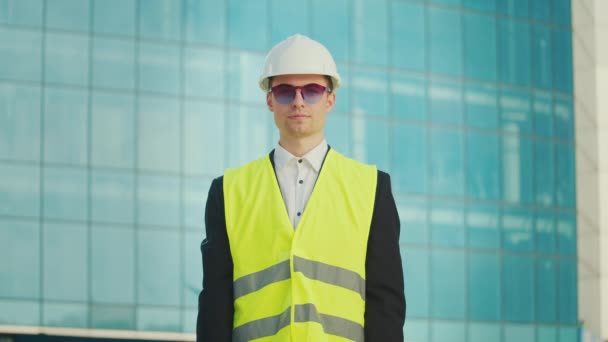 一名成功的年轻男性工程师或穿着头盔和安全夹克的建筑师的画像 他拿着站在蓝窗大楼附近的文件 — 图库视频影像