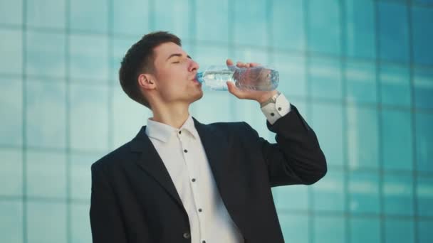 正式なスーツのビジネスマンは 青い窓のある建物の近くに立っている間 ボトルから水を飲む — ストック動画
