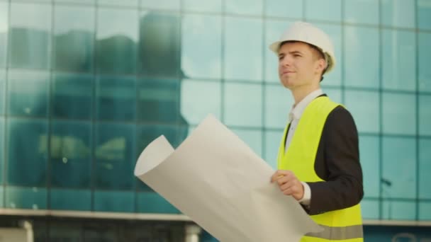 身穿护目镜和安全夹克的男性工程师或建筑师 手拿着施工图纸走在蓝窗大楼附近 — 图库视频影像