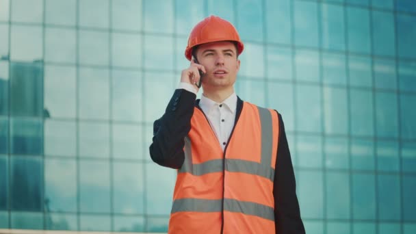 男性工程师或建筑师身穿橙色头盔 身穿安全夹克 手持文件 站在蓝色窗户大楼附近 在电话交谈 — 图库视频影像