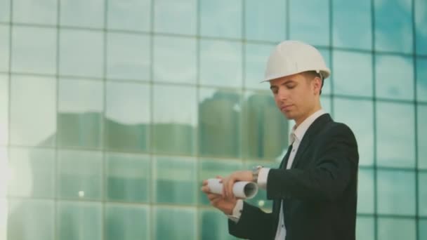 男工程师或建筑师戴着头盔 手拿着建筑图纸走在蓝窗大楼附近 — 图库视频影像