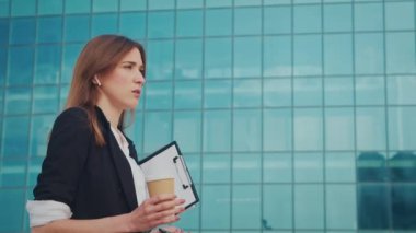 İş kadını kulaklıklı resmi bir takım elbise giyiyor ve modern bir cam iş bölgesinin yakınında yürürken elinde kahve fincanı tutarak telefonla konuşuyor. Kahve molası.