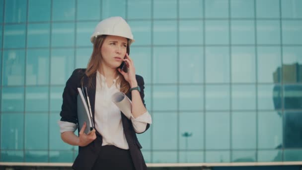 若い女性エンジニアまたはフォーマルスーツとヘルメットスタンディングブルーウィンドウの構造のブループリントを保持し 電話で話す — ストック動画