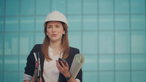 一位身穿正装 头戴耳机的年轻女工程师或建筑师站在蓝窗大楼附近 手持建筑和文件蓝图的电话交谈 — 图库视频影像