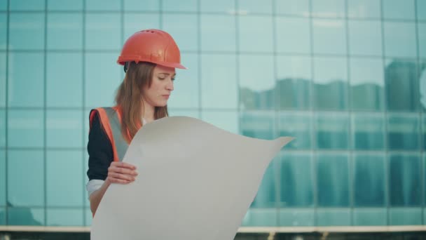 成功した若い女性エンジニアや建築家の肖像オレンジヘルメットと安全ジャケットを着て 青い窓のある建物の近くに立つ彼の手に建設図面を見る — ストック動画