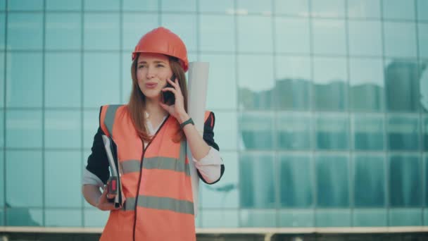 一位年轻的女工程师或建筑师 穿着橙色的头盔和安全衣 站在蓝色窗户的大楼旁边 手握建筑蓝图 在电话上交谈 — 图库视频影像