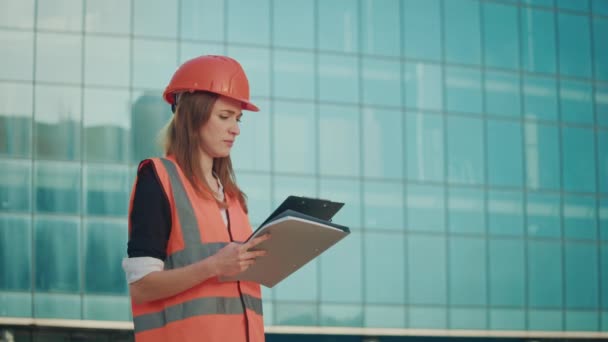 若い女性エンジニアまたは建築家 オレンジヘルメットと安全ジャケット クリップボードを読む — ストック動画