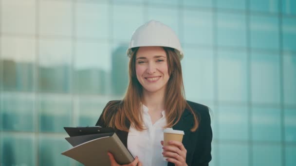 一位成功的年轻女工程师或建筑师头戴白色头盔 手持证件 站在蓝窗大楼旁边喝咖啡的画像 — 图库视频影像